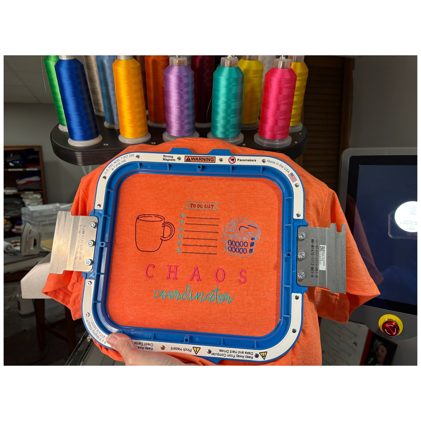Embroidery machine hoop with orange fabric and 'CHAOS coordinator' text, surrounded by spools of thread.