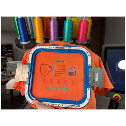 Embroidery machine hoop with orange fabric and 'CHAOS coordinator' text, surrounded by spools of thread.